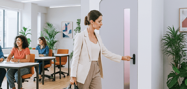 Fotografia que muestra a una mamá que trabaja en un entorno de oficinas acercyose a una cabina de lactancia, al fondo se pueden apreciar dos mujeres trabajyo en sus escritorios respectivamente